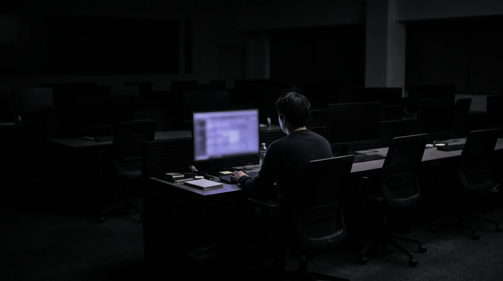 One person seen from behind at the single lit workstation in an otherwise dark, empty office.